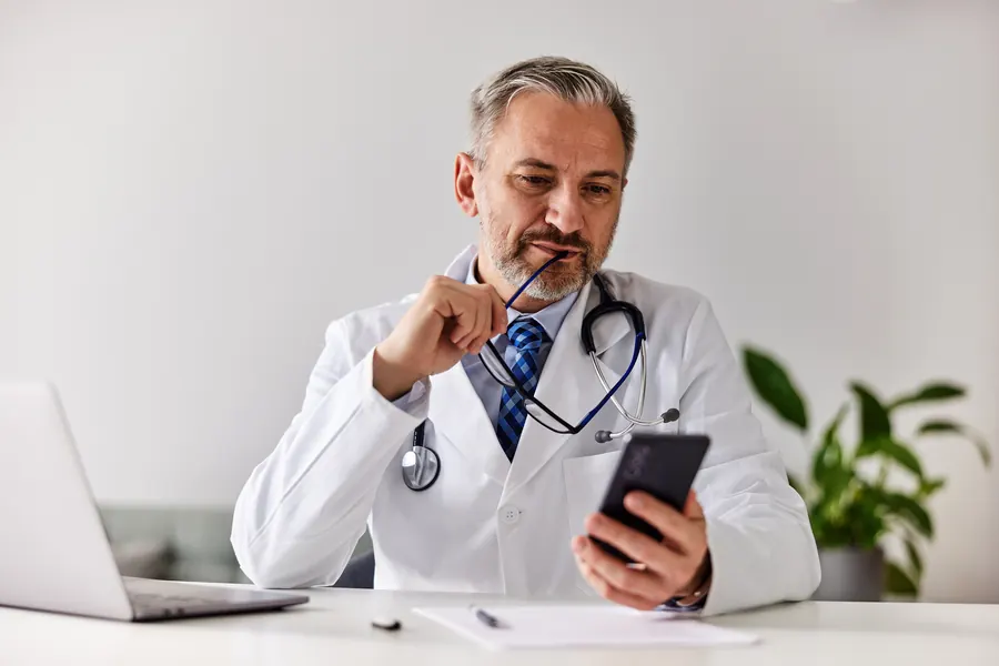 Un médecin en blouse blanche consulte son téléphone à son bureau, l'air pensif