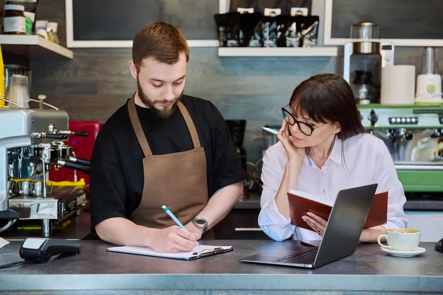 Deux personnes préparent un projet autour d'un carnet et d'un ordinateur, au comptoir d'un café