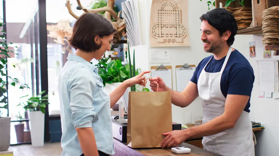 Un commerçant tend son sac de commande à une cliente par-dessus le comptoir : avec le click and collect, la commande est prête avant que le client arrive.