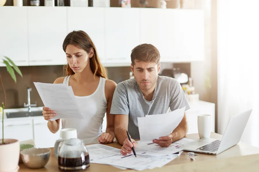 Un couple compare deux documents à la table de la cuisine, un ordinateur portable ouvert à côté