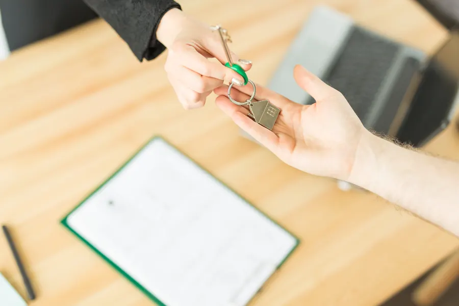 Une main remet une clé à une autre au-dessus d'un bureau en bois, près d'un ordinateur portable