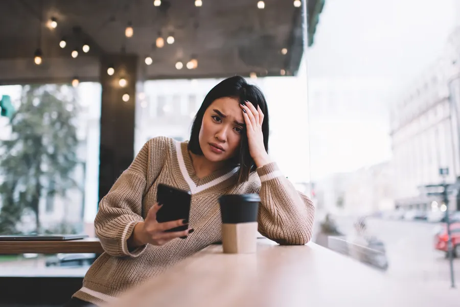 Une femme fixe son téléphone, la main sur le front, à une table de café : conteneurs imbriqués, scripts et add-ons en trop se paient en secondes d’attente.