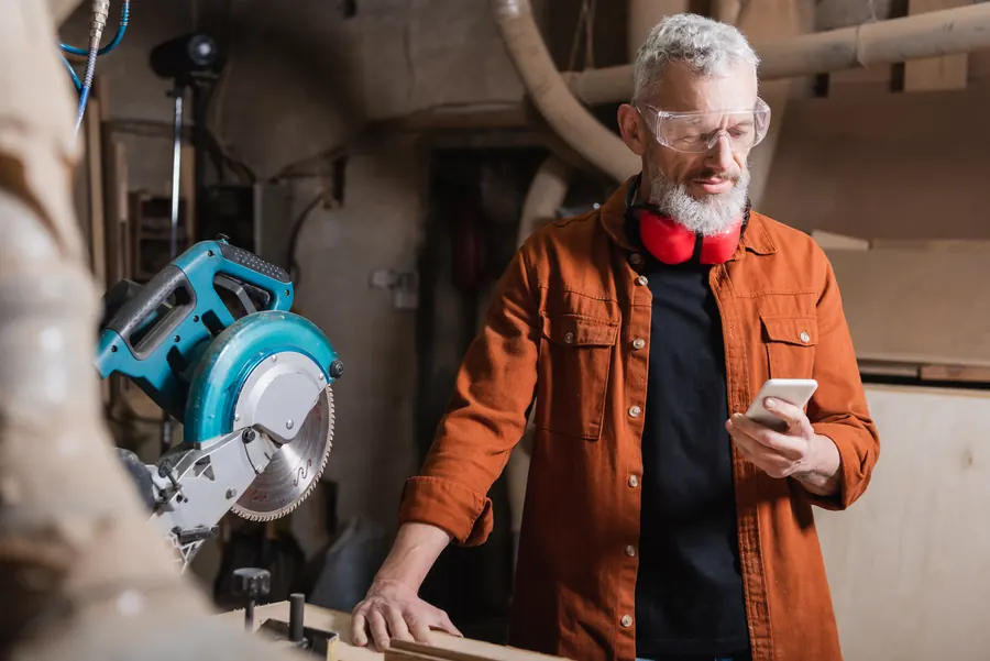 Un menuisier consulte son téléphone à l’atelier : une fois l’envoi fiabilisé, les demandes de devis arrivent là où il les attend.