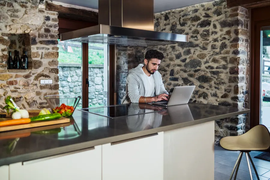 Un propriétaire travaille sur son ordinateur dans la cuisine en pierre de son gîte : relire les conditions des plateformes passe avant tout.