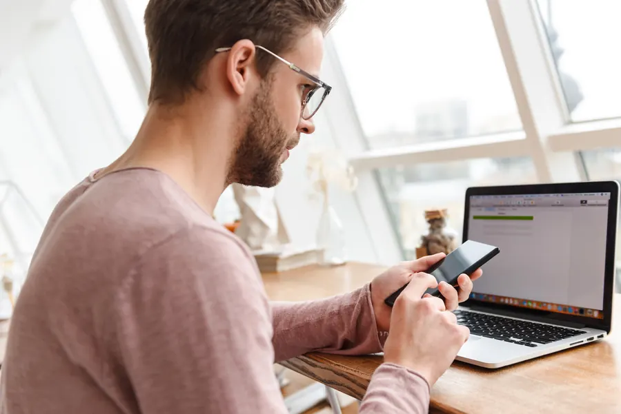 Un homme vérifie un site sur son téléphone et sur son ordinateur : une maintenance sérieuse teste chaque mise à jour avant de la déployer.