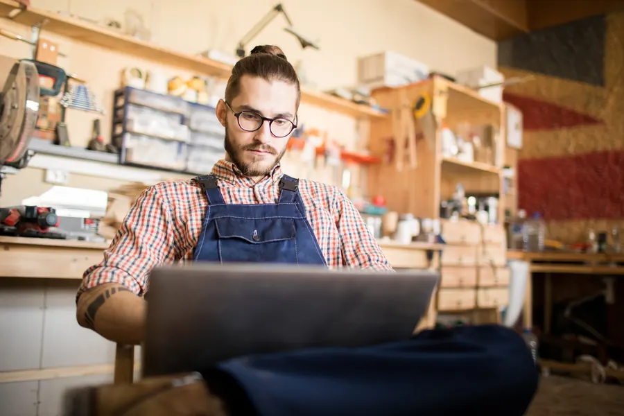 Un artisan en tablier travaille sur son ordinateur portable au milieu de son atelier
