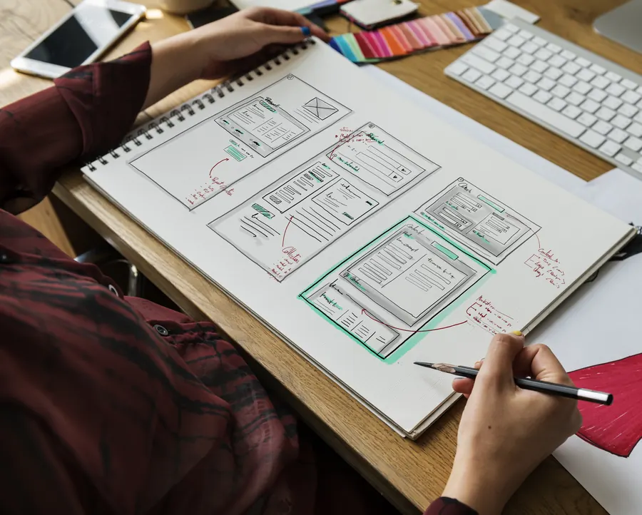 Des croquis de pages de site dessinés au crayon sur un grand carnet, sur un bureau en bois
