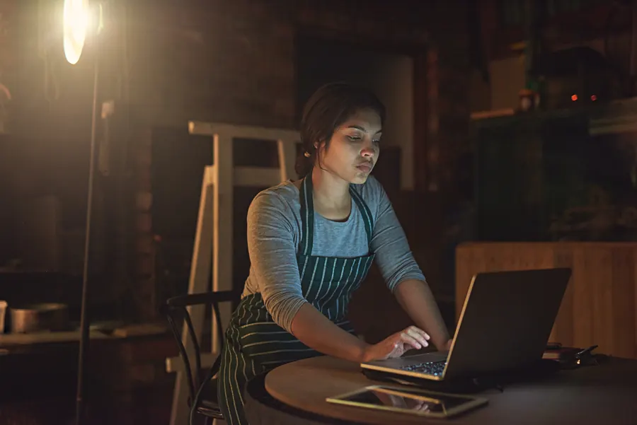 Une artisane devant son ordinateur, tard dans l’atelier : sans maintenance prévue, c’est elle qui gère la panne, souvent au pire moment.