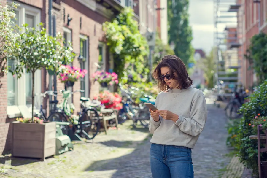 Une femme consulte son téléphone dans une petite rue fleurie, des vélos garés le long des façades