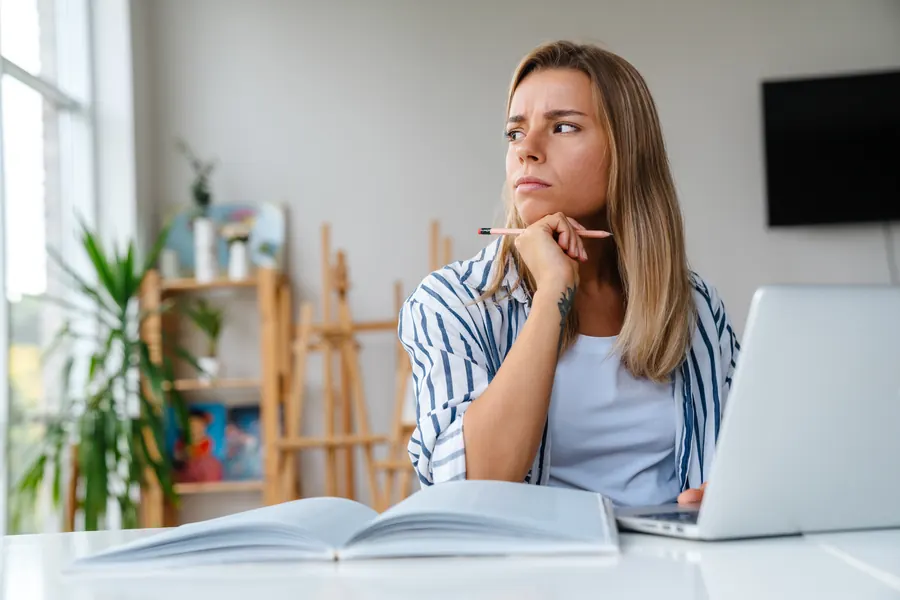 Une femme réfléchit devant son ordinateur portable, à un bureau baigné de lumière naturelle