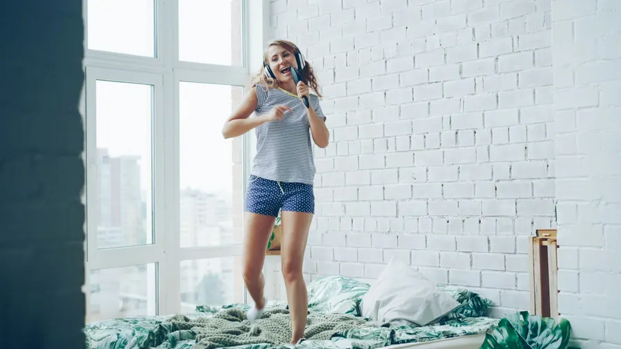 Jeune femme qui danse sur le lit, téléphone à la main, pièce lumineuse