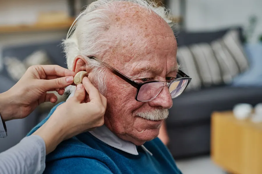 Une audioprothésiste ajuste un appareil sur l'oreille d'un homme de 75 ans, sourires, lumière chaude
