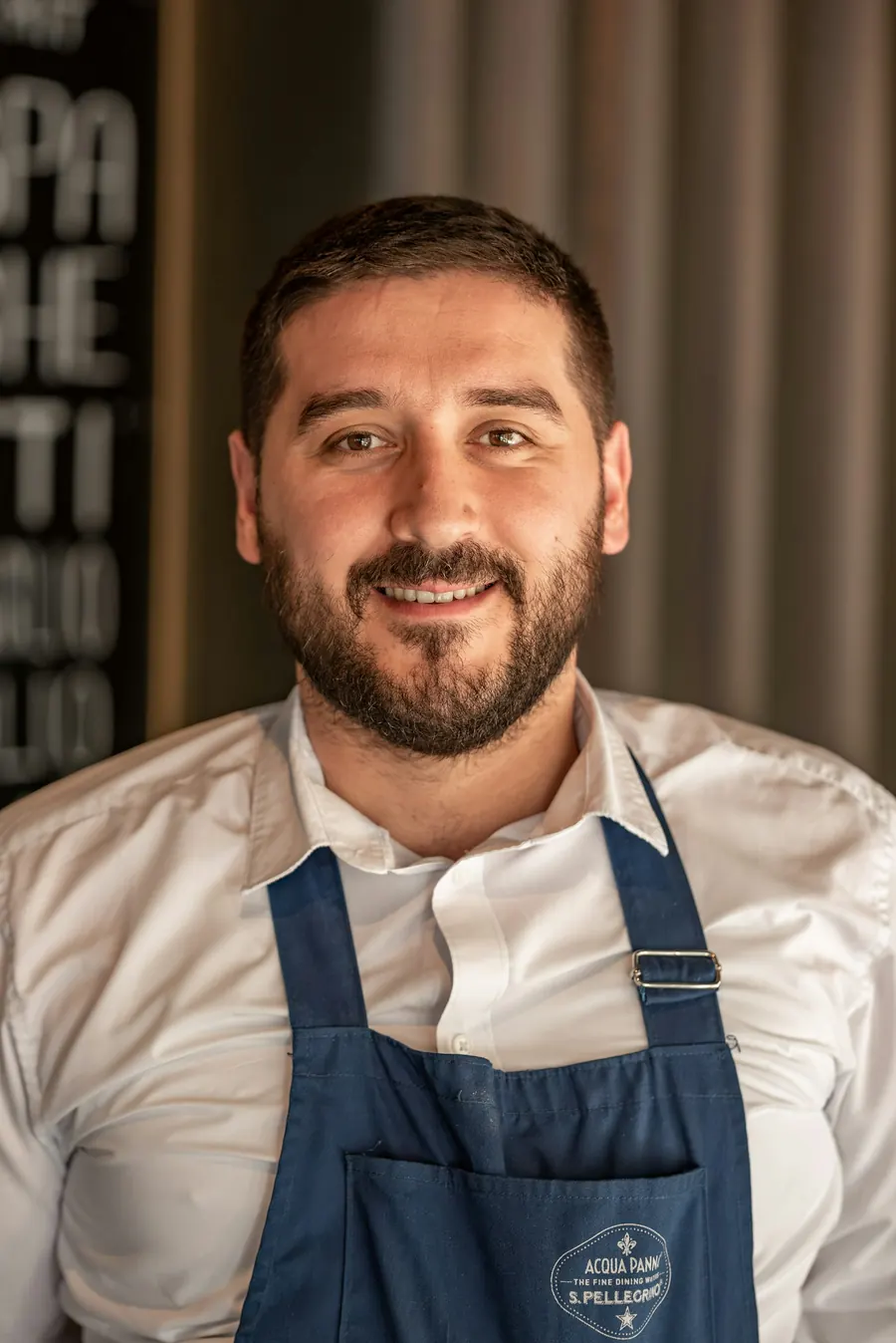 Boulanger d'une quarantaine d'années, tablier fariné, sourire, fournil derrière