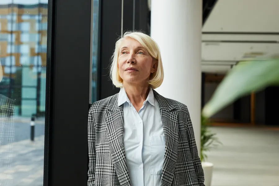 Hélène debout près d'une baie vitrée qui donne sur la ville, lumière du matin, de trois quarts