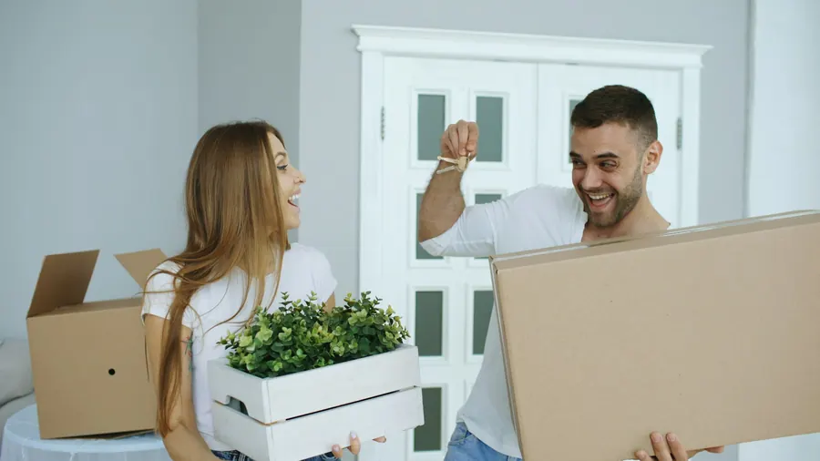 Couple souriant qui emménage avec des cartons, geste de la main en l'air