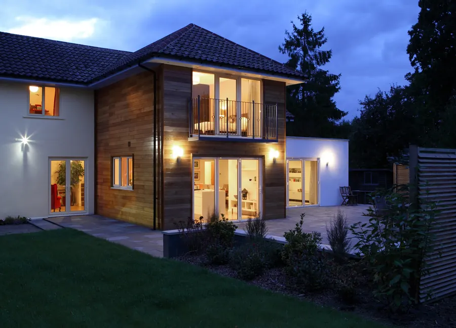 Maison neuve contemporaine au crépuscule, fenêtres éclairées, toit de tuiles, bardage bois, pelouse jeune