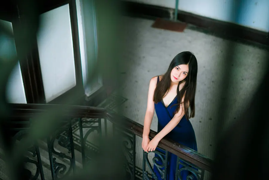 Jeune femme en robe bleu nuit accoudée à la rampe en fer forgé d'un escalier ancien