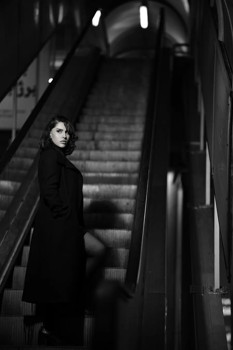 Femme en manteau noir sur un escalier mécanique, photo en noir et blanc