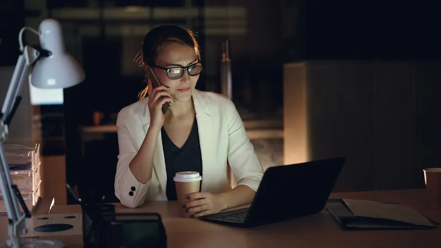 Intervenante au téléphone devant un ordinateur portable, bureau calme en soirée, lampe allumée