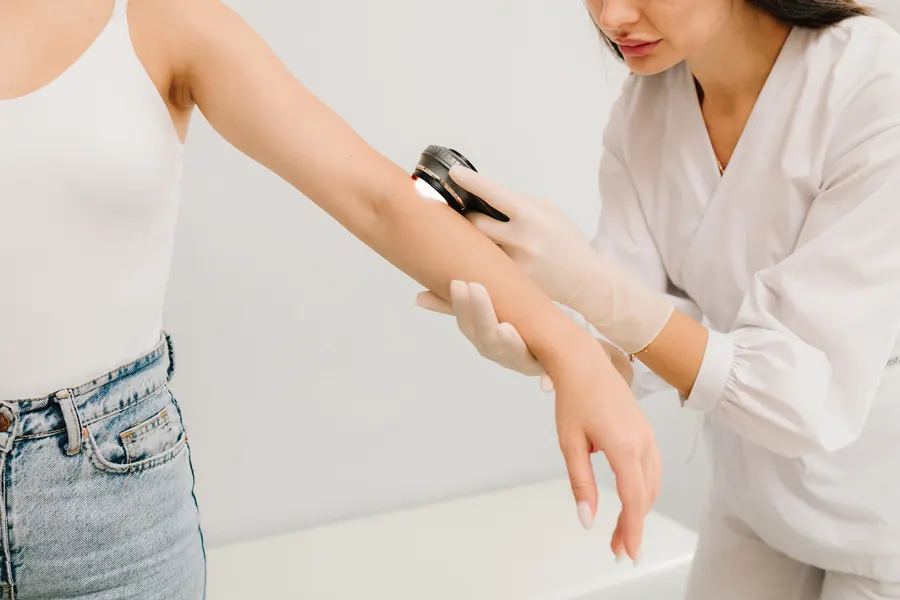 Dermatologue qui examine l'avant-bras d'une patiente au dermatoscope, cabinet clair et épuré, visage de la patiente peu visible