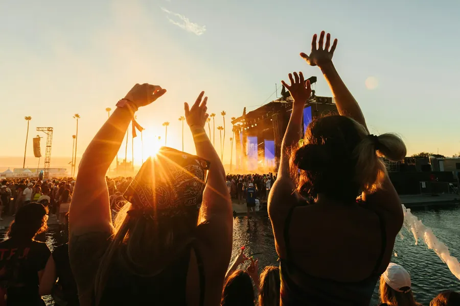 Festivaliers bras levés face au soleil couchant, près d'une scène au bord de l'eau
