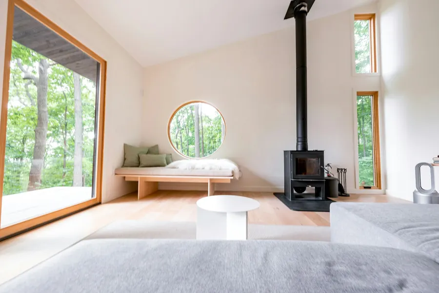 Intérieur de cabane au matin, petit poêle à bois, lit sous une couverture de laine, fenêtre sur les branches