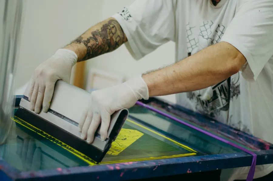 Sérigraphe qui tire la racle sur un carrousel à t-shirts, encre jaune, atelier