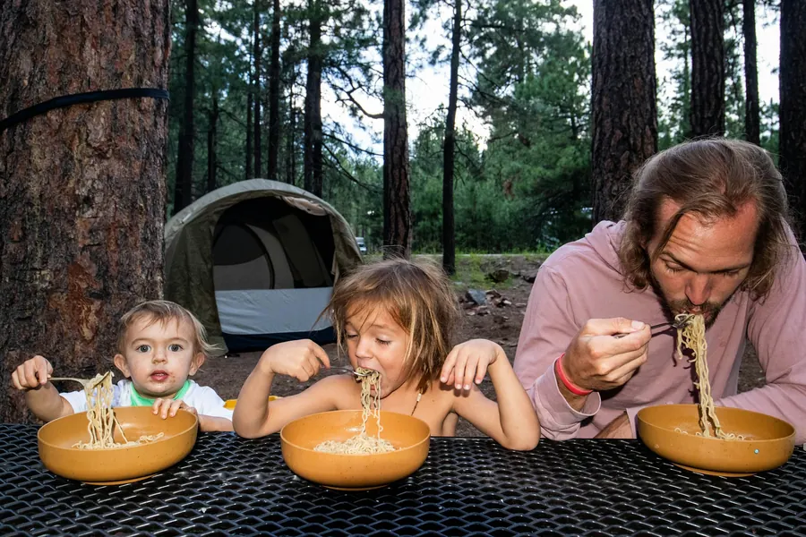 Un père et deux enfants mangent en camping, sans camping-car visible