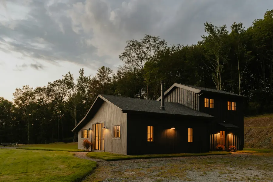 Maison à bardage bois sombre aux fenêtres éclairées, au crépuscule, en lisière de forêt
