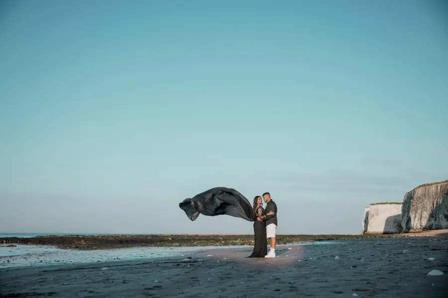 Mariés sur une plage de galets, tissu sombre au vent, ciel clair
