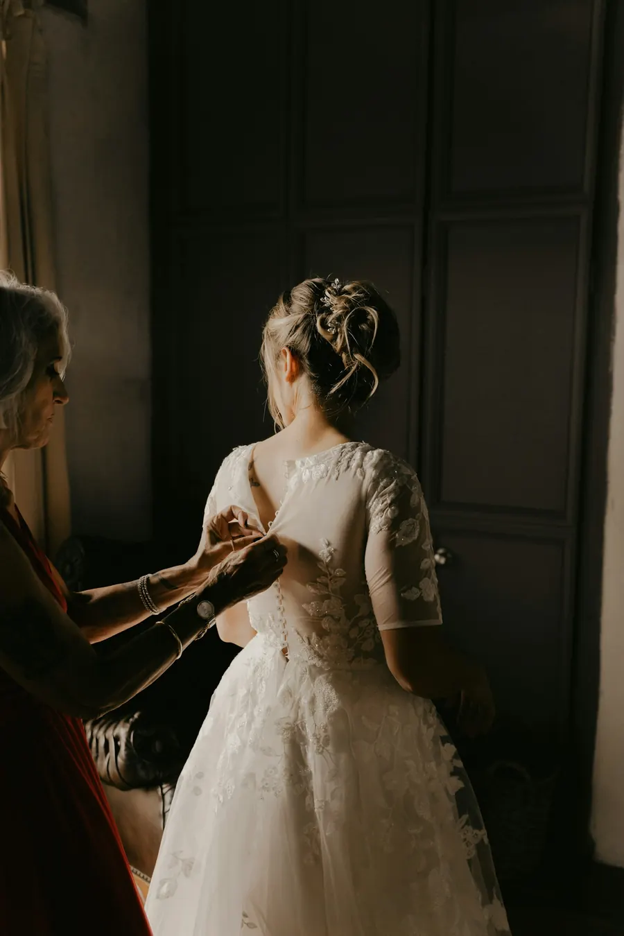 Mère qui boutonne la robe de la mariée près d'une fenêtre, lumière naturelle