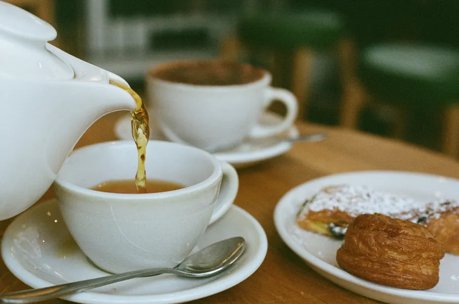 Thé versé dans une tasse en porcelaine, part de gâteau à côté, table en bois, lumière de fenêtre