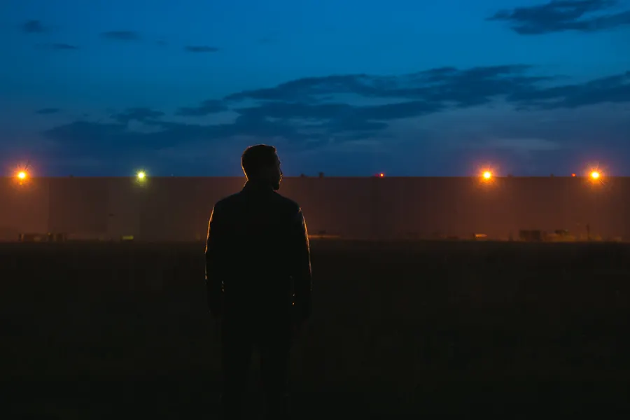 Agent en blouson noir sans insigne, lampe à la main, devant un bâtiment industriel au crépuscule