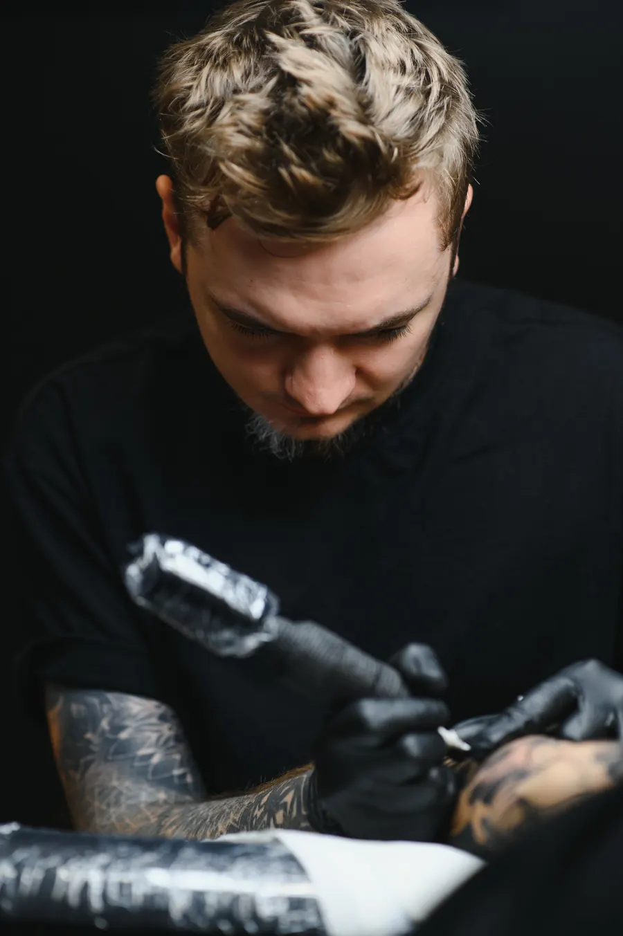 Tatoueur en gants noirs qui tatoue un avant-bras, lumière de lampe, fond sombre