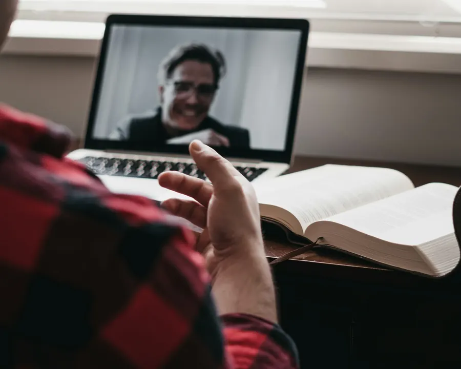 Personne en visioconférence sur ordinateur portable, livre ouvert à côté