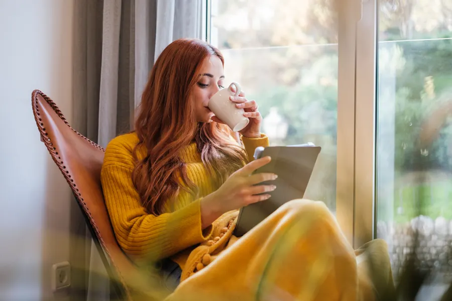 Femme lisant sur une tablette, une tasse à la main, près d’une fenêtre