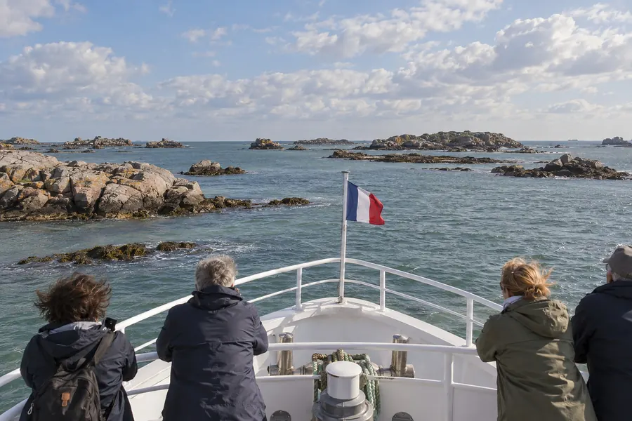 Passagers de dos sur le pont avant d’une vedette, face aux îlots de granit de l’archipel, pavillon français à la proue