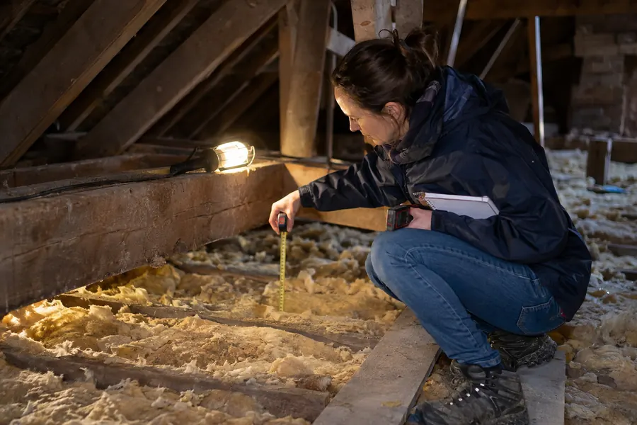 Maud Lecoeur, accroupie sur une planche dans des combles perdus, mesure au mètre ruban l’épaisseur d’une laine ancienne tassée et jaunie entre les solives, éclairée par une lampe baladeuse