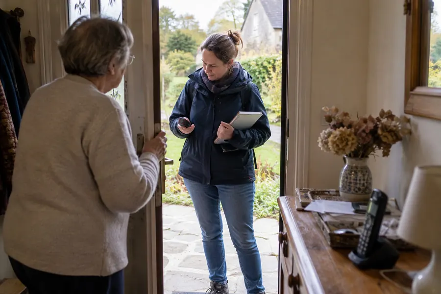 Une dame âgée en gilet clair tient sa porte d’entrée ouverte ; sur le seuil, Maud Lecoeur, carnet à la main, lui parle. Au premier plan, sur la console de l’entrée, un téléphone sans fil et un vase de fleurs séchées