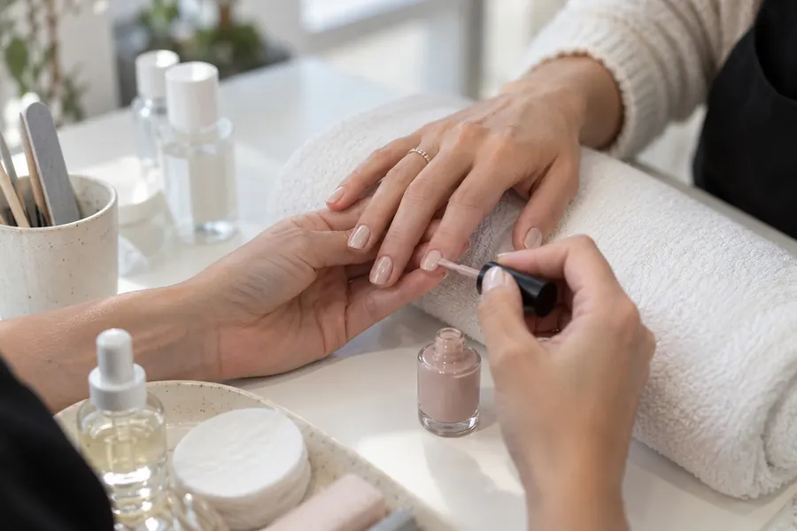 Manucure : l’esthéticienne pose un vernis nude sur les ongles d’une cliente dont la main repose sur une serviette roulée, flacons et cotons sur la table blanche