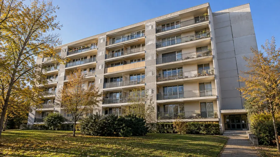Une résidence de six étages des années 1970 à balcons filants et panneaux de béton clair, devant une pelouse et des arbres aux feuilles jaunies, sous un ciel bleu d’automne
