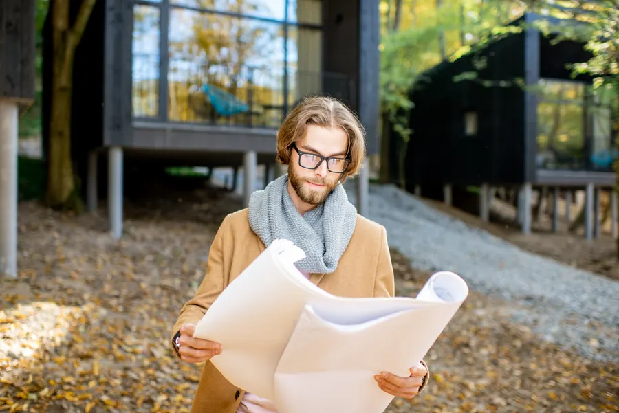 Un homme à lunettes, manteau camel et écharpe grise, déroule un grand plan papier devant des maisons à ossature posées sur pilotis, au milieu des feuilles d’automne